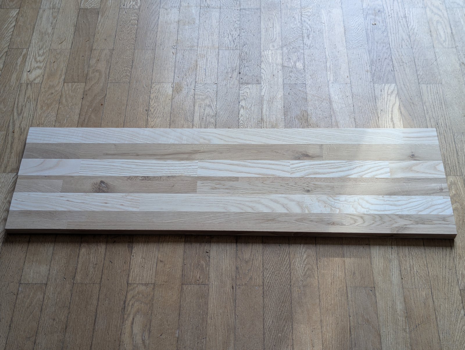 Long panneau rectangulaire formé de bandes de bois claires et foncées, posé sur le sol.