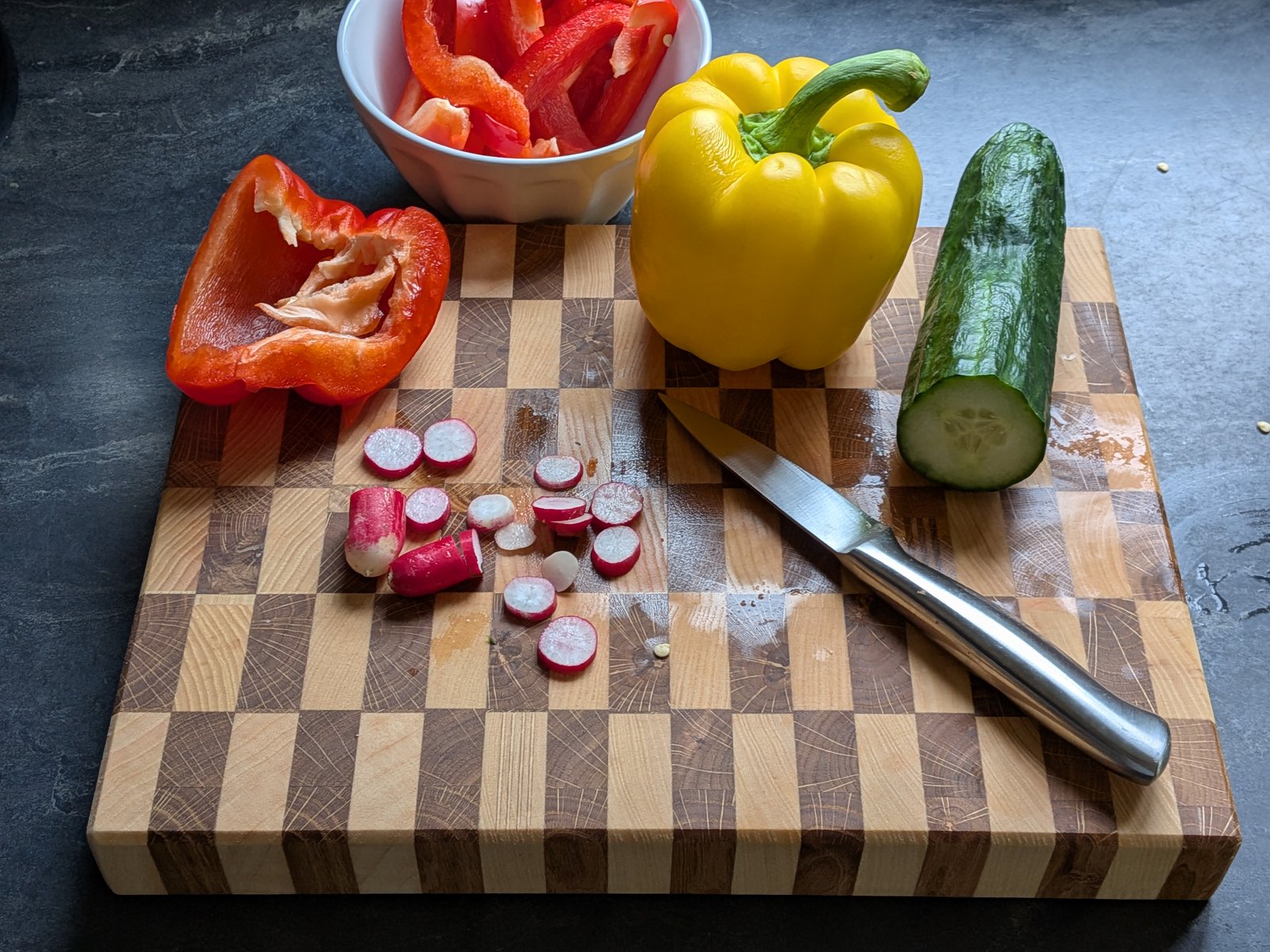 Planche en damier portant des radis coupés, des poivrons rouge et jaune, un concombre et un couteau.